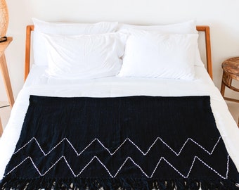 May include: A black throw blanket with a white zigzag pattern and fringe detail is displayed on a bed with white linens. The bed has a wooden frame and is in a bright room with a wooden side table and a woven lamp.