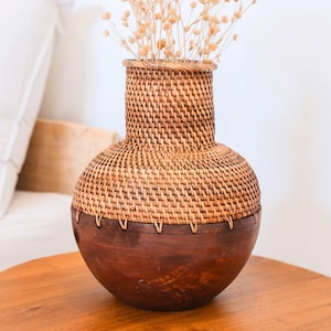 Könnte beinhalten: Eine dekorative Vase mit einem gewebten Rattantop und einem dunkelbraunen Holzsockel. Die Vase ist mit getrockneten, beigefarbenen Blumen gefüllt. Die Vase steht auf einem runden Holztisch.