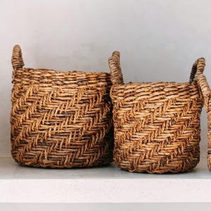 May include: Three woven baskets with handles. The baskets are made of natural-colored material and have a textured, diagonal weave pattern. The baskets are different sizes and are arranged side by side.