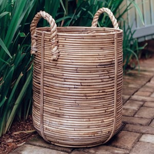 Puede incluir: Una cesta de mimbre tejida con asas de cuerda. La cesta es de color marrón claro y tiene un aspecto natural y rústico. Está sentada en un patio de adoquines.