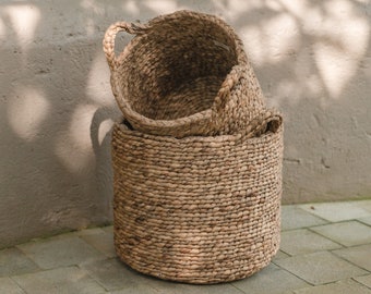 Peut inclure: Deux paniers tissés en matériaux naturels. Le panier du haut est rond avec deux poignées et le panier du bas est cylindrique. Les paniers sont posés sur un sol en carrelage brun clair.