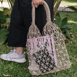 Può includere: Una borsa tote in macramè beige con un design a rete intrecciata, che contiene un libro rosa e nero. La borsa ha manici spessi in corda e viene trasportata su pantaloni neri e scarpe da ginnastica grigie su uno sfondo erboso.