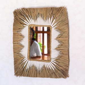 Peut inclure: Miroir rectangulaire avec un cadre tissé naturel. Le cadre présente un motif d'éléments blancs et beiges rayonnant du bord du miroir. Le miroir reflète une personne en chemise blanche et une fenêtre.