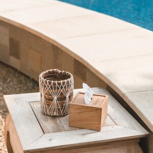 May include: A wooden tissue box with a white tissue inside, sitting on a light wood table next to a woven basket with a white rope design.