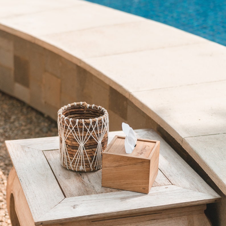 May include: A wooden tissue box with a white tissue inside, sitting on a light wood table next to a woven basket with a white rope design.