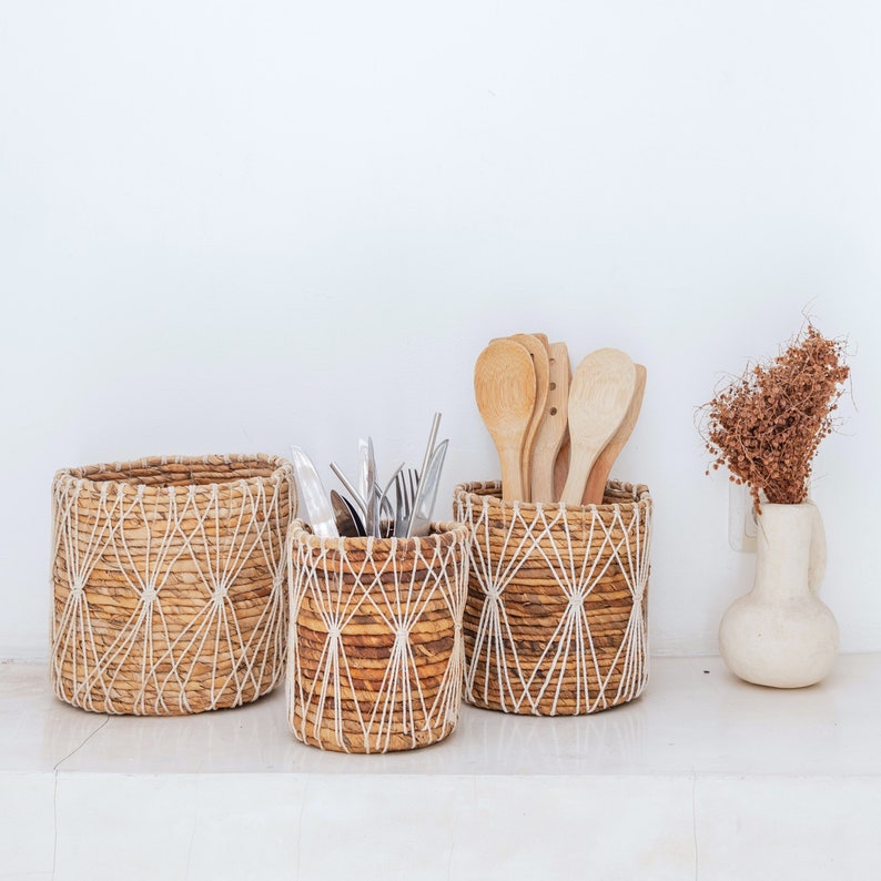 May include: Three woven baskets with a natural, light brown color. The baskets are made of a woven material with a white string pattern. The baskets are sitting on a white surface. The basket on the left is empty. The middle basket is filled with silverware. The basket on the right is filled with wooden spoons.