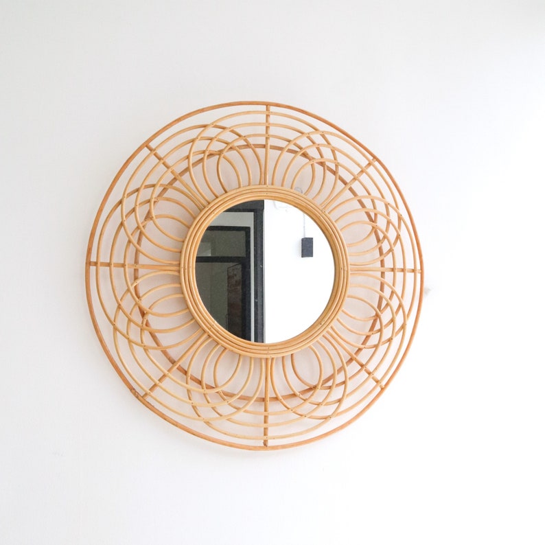 May include: A round, woven rattan mirror with a natural wood finish. The mirror has a circular frame with a floral design. The mirror is reflecting a room with a white wall and a door.
