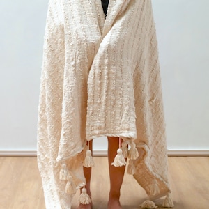 May include: A cream-colored throw blanket with a textured weave and fringe tassels. The blanket is draped over a person's shoulders.