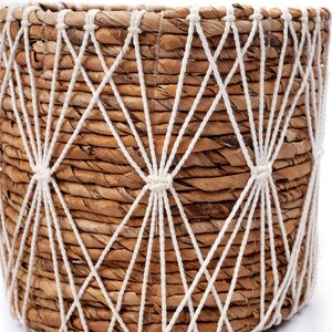 May include: A woven seagrass planter with a white rope design. The planter has a natural brown color and a round shape.