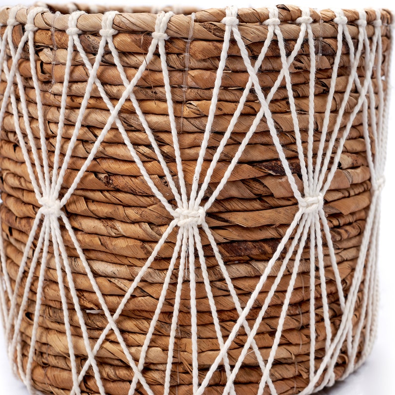 May include: A woven seagrass planter with a white rope design. The planter has a natural brown color and a round shape.