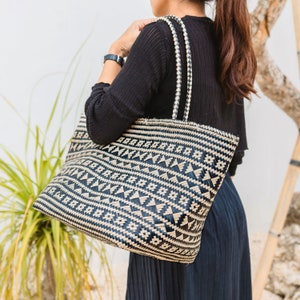 May include: A black and white woven tote bag with a geometric pattern. The bag has two handles and is being held by a person wearing a black shirt and a black skirt.