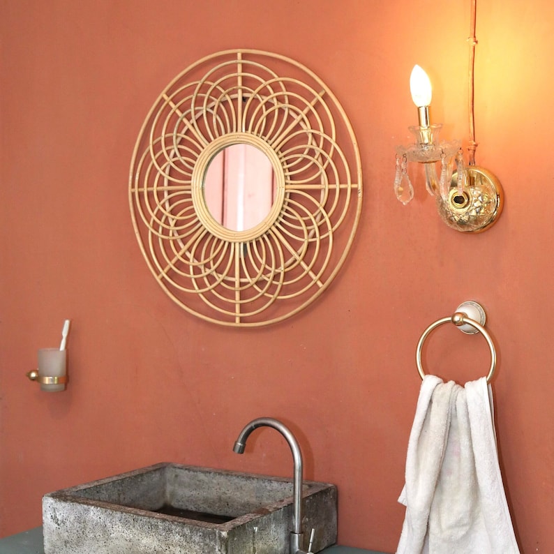 May include: A round woven rattan mirror with a circular mirror inside, hanging on a peach wall with a gold-toned light fixture and a towel rack with a white towel hanging on it.