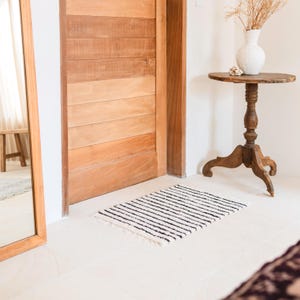 May include: A black and white striped rug with a fringed edge is placed in front of a wooden door. The rug is made of a soft, fluffy material.