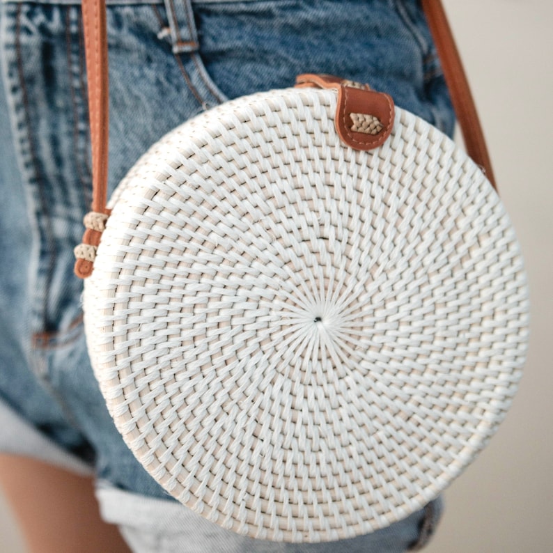 May include: White round woven straw bag with a brown leather strap. The bag has a circular woven pattern.