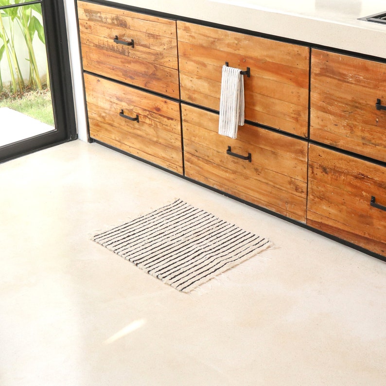 May include: A small, rectangular, cream-colored rug with black stripes. The rug is placed in front of a set of wooden cabinets with black handles.