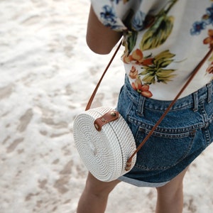 May include: A white round woven bag with a brown leather strap and a gold buckle. The bag is worn as a crossbody bag.