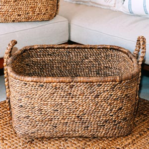 Puede incluir: Una cesta rectangular tejida con asas trenzadas. La cesta está hecha de fibras de color natural y se encuentra sobre una estera tejida a juego. La cesta tiene forma cuadrada y está diseñada para el almacenamiento.