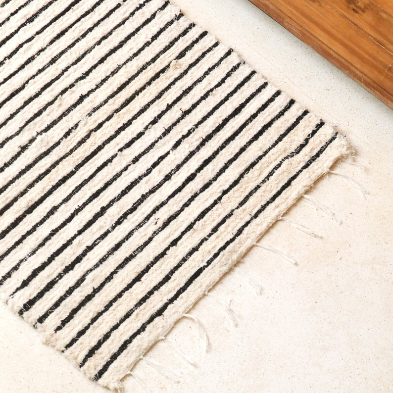 May include: A white and black striped rug with a frayed edge. The rug is made of a natural fiber and has a simple, minimalist design.