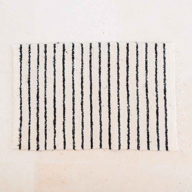 May include: A white and black striped rug with a woven texture. The rug has a frayed edge.