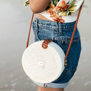 May include: A white round woven bag with a brown leather strap. The bag is worn over the shoulder of a person wearing a floral top and denim shorts.