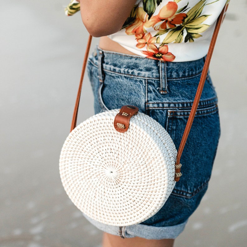 May include: A white round woven bag with a brown leather strap. The bag is worn over the shoulder of a person wearing a floral top and denim shorts.