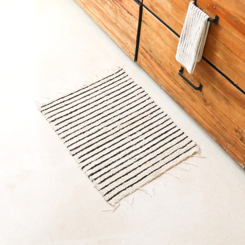 May include: A white and black striped rug with frayed edges. The rug is made of a natural fiber and has a simple, minimalist design.