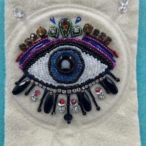 May include: A white felt background with a beaded evil eye design. The eye is blue with black and white beads surrounding it. The eye is surrounded by black beads and black teardrop-shaped beads.
