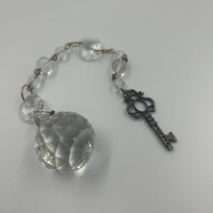 May include: A clear crystal ball and beads are connected by gold-colored metal links. A decorative silver-colored key charm hangs from the chain. The items are displayed on a white surface.