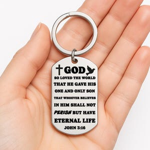 Könnte beinhalten: Ein silberfarbener Schlüsselanhänger mit einem rechteckigen Anhänger. Der Anhänger zeigt den Text "+GOD" und eine Taubengrafik sowie einen Bibelvers. Der Text lautet: "SO LOVED THE WORLD THAT HE GAVE HIS ONE AND ONLY SON THAT WHOEVER BELIEVES IN HIM SHALL NOT PERISH BUT HAVE ETERNAL LIFE JOHN 3:16."