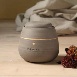 May include: A gray ceramic urn with a gold heart and the name "Fiona" inscribed on it. The urn has a lid and is sitting on a wooden surface.