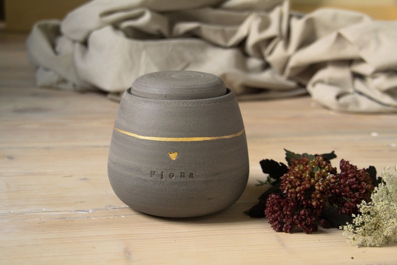 May include: A gray ceramic urn with a gold stripe and a gold heart with the name Fiona inscribed on it. The urn is sitting on a wooden surface with a small bouquet of flowers.
