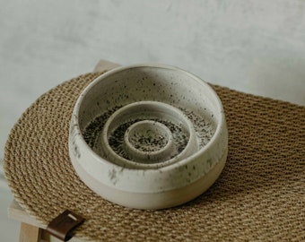 May include: White ceramic dog bowl with a textured finish and three concentric circles. The bowl is on a brown woven mat.