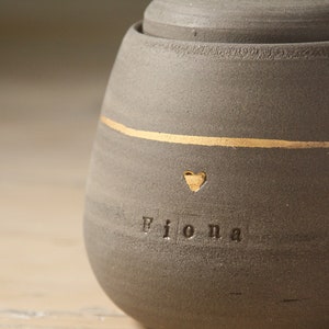 May include: A gray ceramic jar with a gold stripe and the name "Fiona" inscribed on the front. A small heart is above the name.