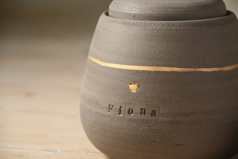 May include: A gray ceramic jar with a gold stripe and the name "Fiona" inscribed on the front. A small heart is above the name.
