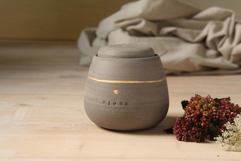 May include: A gray ceramic urn with a gold heart and the name "Fiona" inscribed on it. The urn has a lid and is sitting on a wooden surface.