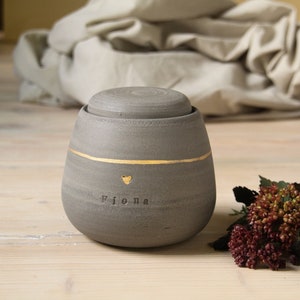 May include: A gray ceramic urn with a gold band and a gold heart. The urn is inscribed with the name "Fiona".