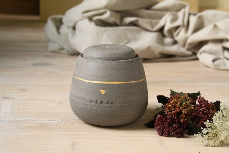 May include: A gray ceramic urn with a gold band and a gold heart. The urn is inscribed with the name "Fiona".
