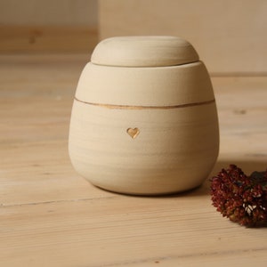 May include: A cream-colored ceramic urn with a gold trim and a heart carved into the front. The urn has a lid and is sitting on a wooden surface.