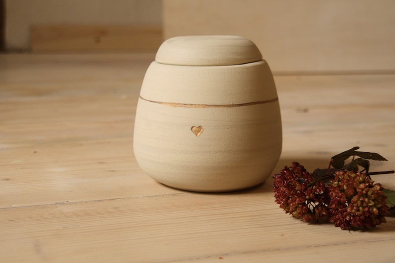 May include: A cream-colored ceramic urn with a gold trim and a heart carved into the front. The urn has a lid and is sitting on a wooden surface.