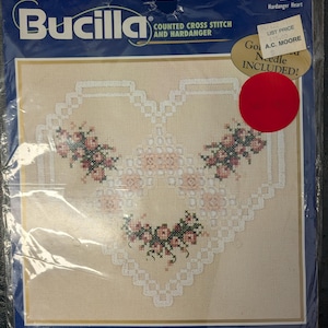 May include: A Bucilla Hardanger Heart counted cross stitch kit in a plastic package. The kit features a heart-shaped design with white geometric patterns and pink floral accents on a tan fabric. The package includes text and an 8.5 inch by 8 inch picture.