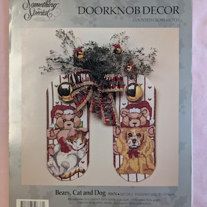 May include: A counted cross stitch pattern for a door knob decoration featuring a bear, cat, and dog wearing Santa hats. The pattern is titled "Bears, Cat and Dog" and is part of a set of two. The finished size is 5 inches by 12 inches.