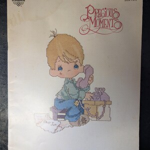 May include: A cross-stitch pattern featuring a crying cartoon boy holding a telephone. The design includes the text "Precious Moments" and "Dear Jon". The pattern is on a cream-colored background.