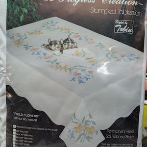 May include: A packaged white tablecloth with a floral cross-stitch design, labeled "Field Flowers." The package includes a variety of size options, including square, oblong, and oval. The text "A Progress Creation" is visible.