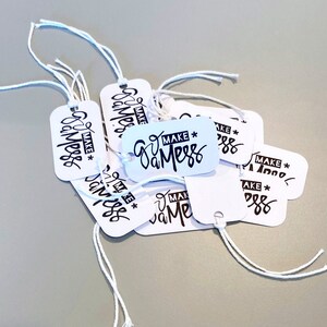 Puede incluir: Un montón de etiquetas de regalo blancas con un cordón negro atado. Cada etiqueta tiene el texto "MAKE a MESS" escrito a mano.