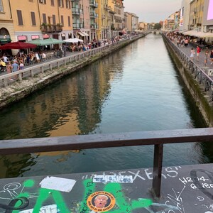 May include: A view of a canal in a city with buildings on either side. The canal is lined with people walking along the edge. There are several stickers on a railing, including one with a picture of Donald Trump. The text on the sticker reads "NEW CAN SO MUCH BUT COME OUT OF ONE ASSHOLE."