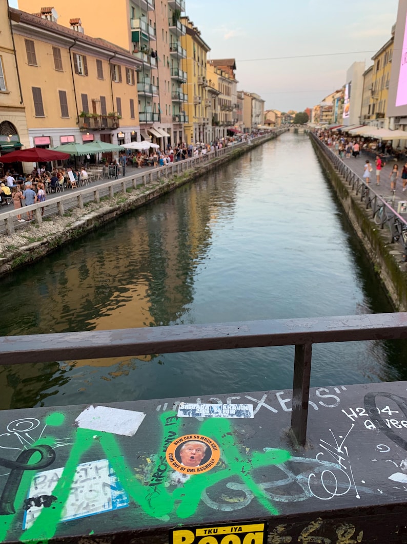 May include: A view of a canal in a city with buildings on either side. The canal is lined with people walking along the edge. There are several stickers on a railing, including one with a picture of Donald Trump. The text on the sticker reads "NEW CAN SO MUCH BUT COME OUT OF ONE ASSHOLE."
