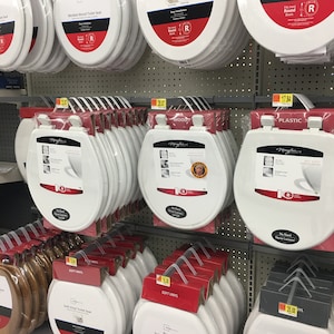 May include: A display of toilet seats in various materials, including wood, plastic, and soft vinyl. The seats are stacked and labeled with their material and price. The seats are all white and have a round shape.