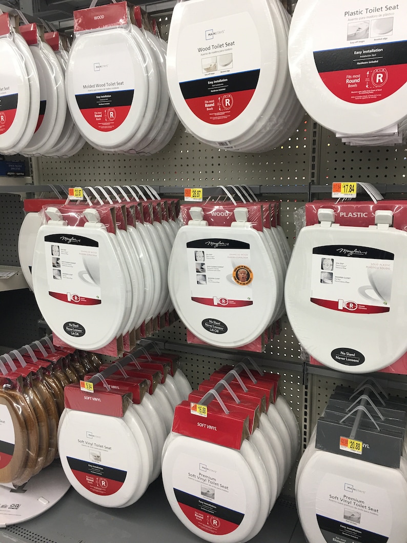 May include: A display of toilet seats in various materials, including wood, plastic, and soft vinyl. The seats are stacked and labeled with their material and price. The seats are all white and have a round shape.