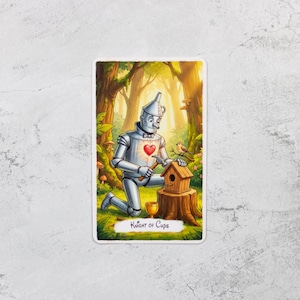 May include: A tarot card illustration featuring the Tin Man from The Wizard of Oz, kneeling and hammering a birdhouse. The card is labeled "Knight of Cups" and set in a forest scene with a bird perched nearby. The Tin Man has a glowing heart.