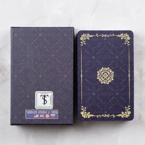Pu&ograve; includere: Un mazzo di tarocchi e un set di scatole. La scatola e i dorsi delle carte sono blu scuro con un motivo ripetuto. I dorsi delle carte hanno accenti floreali dorati e un emblema centrale. La scatola ha un adesivo che recita "TAROCCO STUDIO &copy; 2026".